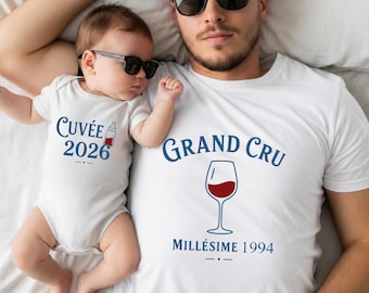 Può includere: Una t-shirt bianca a maniche corte con il testo GRAND CRU e MILLÉSIME 1994 sopra e sotto un grafico di un calice di vino, e un body bianco per neonati con CUVÉE 2026 e un biberon, entrambi con scritte blu.