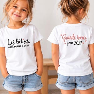 Peut inclure: Un t-shirt blanc à manches courtes avec l'inscription noire Les bêtises, c'est mieux à deux... sur le devant et le texte rose Grande soeur pour 2027 accompagné de petits cœurs roses imprimés au dos du vêtement.