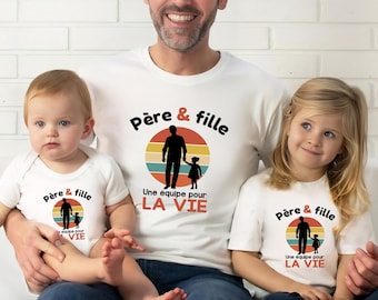 Op de afbeelding: Een wit t-shirt en een witte baby-romper met een afbeelding van het silhouet van een vader en dochter in een zonsondergangcirkel. De tekst luidt "Père & fille" in het zwart en "Une équipe pour LA VIE" in zwarte en oranje letters.