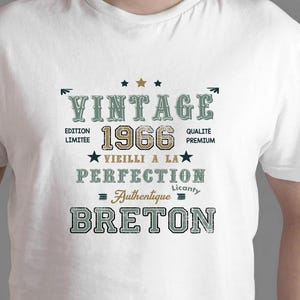 Peut inclure: T-shirt blanc avec un design vintage. Le design comprend le texte "VINTAGE 1966", "BRETON" et d'autres phrases françaises. Le texte est dans différentes polices et couleurs, dont le vert, le marron et le noir. Le t-shirt a un col rond.