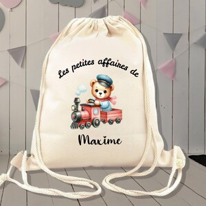 Op de afbeelding: Een witte trekkoordtas met de tekst "Les petites affaires de Maxime" en een cartoonillustratie van een teddybeer die een rode speelgoedtrein bestuurt.