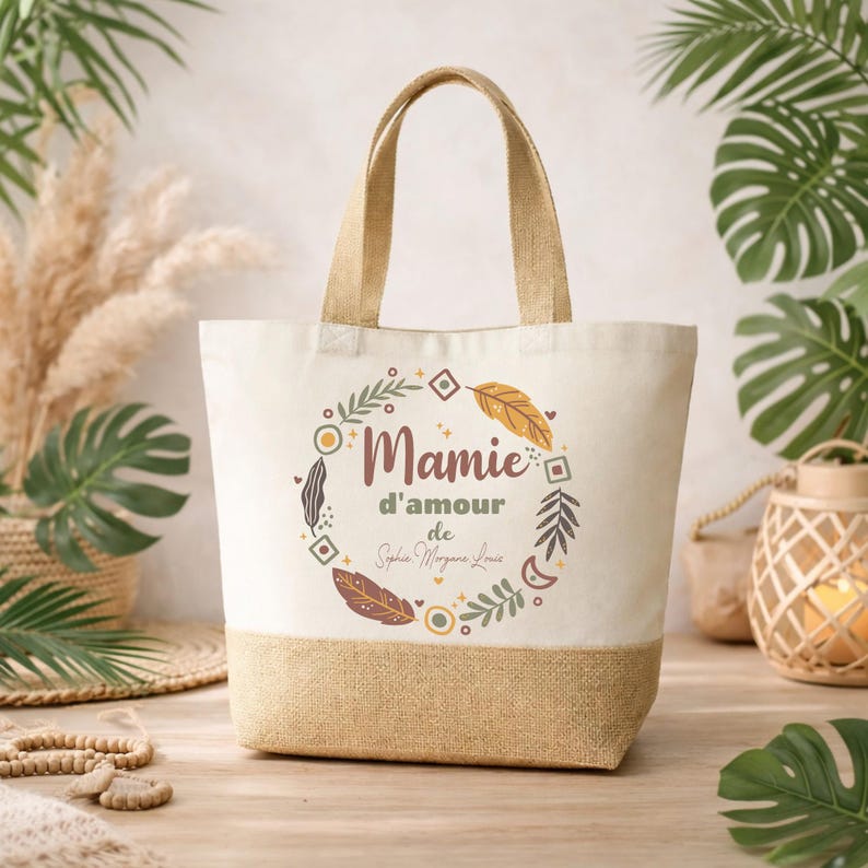 Puede incluir: Un bolso tote beige con base y asas de yute natural. El bolso presenta las palabras "Mamie d'amour" y un nombre dentro de un dise&ntilde;o circular de hojas y plumas. El bolso est&aacute; sobre una superficie de madera.