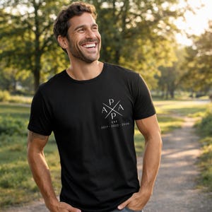 May include: Black crew neck t-shirt with a white graphic on the chest. The graphic features the letters "P A P A" in a cross design, with "EST 2017-2026" below. The shirt is worn outdoors.