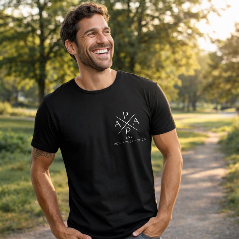 May include: Black crew neck t-shirt with a white graphic on the chest. The graphic features the letters "P A P A" in a cross design, with "EST 2017-2026" below. The shirt is worn outdoors.