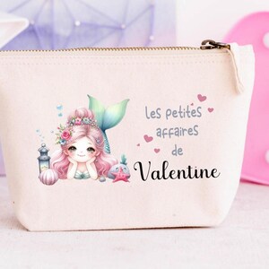 Peut inclure: Une trousse crème zippée avec une illustration de sirène aux cheveux roses et à la queue turquoise. Le texte "Les petites affaires de Valentine" est imprimé sur la trousse, avec de petits cœurs roses. Une lumière décorative rose en forme de cœur est en arrière-plan.