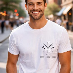May include: White t-shirt with a black graphic design on the front. The design includes the letters "P", "A", and "EST" with the years 2017, 2022, and 2026. The shirt is worn by a person smiling in an outdoor setting.