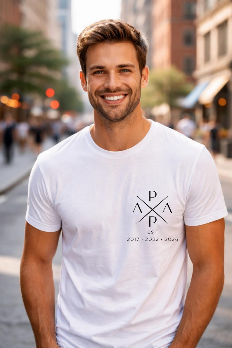 May include: White t-shirt with a black graphic design on the front. The design includes the letters "P", "A", and "EST" with the years 2017, 2022, and 2026. The shirt is worn by a person smiling in an outdoor setting.