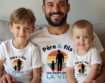 Op de afbeelding: Drie witte t-shirts met een afbeelding van het silhouet van een vader en kind tegen een retro zonsondergangcirkel. De tekst "Père & fils" en "Une équipe pour LA VIE" staat in zwarte en blauwgroene letters op de borst van elk kledingstuk.