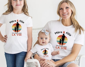 Op de afbeelding: Twee witte T-shirts met korte mouwen en een witte babybody met korte mouwen, elk voorzien van een afbeelding van een moeder en dochter silhouet tegen een retro zonsondergangcirkel, met de tekst Mère & fille Une équipe pour LA VIE.