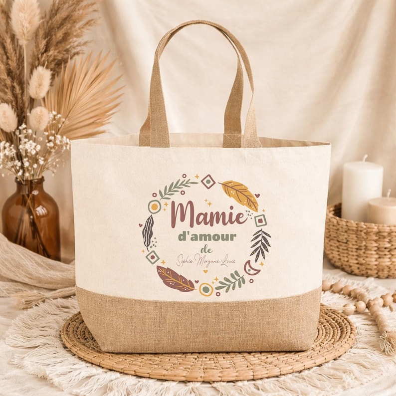 Puede incluir: Un bolso tote beige con base y asas marrones. El bolso presenta un dise&ntilde;o circular con las palabras "Mamie d'amour" y un nombre, rodeado de hojas y plumas. El bolso est&aacute; sobre una esterilla tejida.