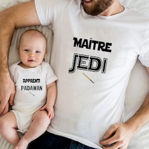 Puede incluir: Camisetas blancas y body de bebé con texto temático de Star Wars. La camiseta de adulto dice "MAÎTRE JEDI" y el body del bebé dice "APPRENTI PADAWAN". Ambos tienen un gráfico de sable de luz amarillo.