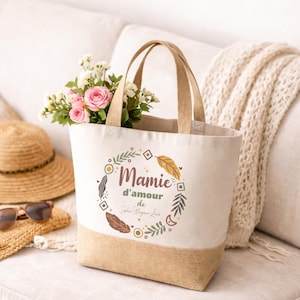 Puede incluir: Un bolso de mano de lona con base y asas beige, lleno de flores rosas y blancas. La bolsa presenta el texto "Mamie d'amour" en un dise&ntilde;o de corona decorativa. Un sombrero de paja y gafas de sol descansan cerca.
