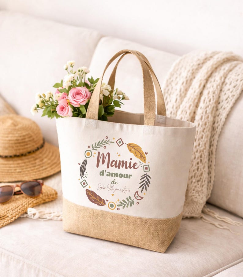 Puede incluir: Un bolso de mano de lona con base y asas beige, lleno de flores rosas y blancas. La bolsa presenta el texto "Mamie d'amour" en un dise&ntilde;o de corona decorativa. Un sombrero de paja y gafas de sol descansan cerca.