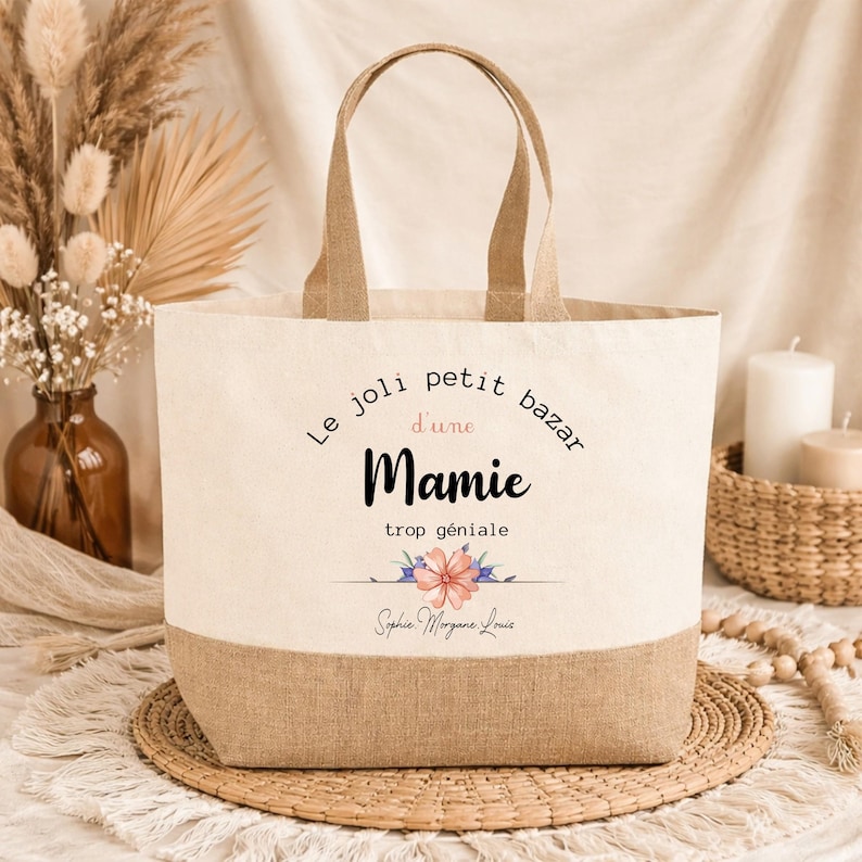 May include: A beige tote bag with a brown bottom and handles. The bag features the text "Le joli petit bazar d'une Mamie trop g&eacute;niale" and a floral design. The bag is made of canvas and burlap.