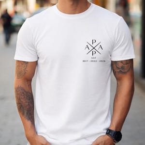 May include: White crew neck t-shirt with a black graphic on the upper left chest. The graphic features the letters P, A, and X arranged in a cross shape, with the text "EST 2017-2022-2026" below.