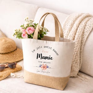 May include: A canvas tote bag with a natural jute bottom and handles. The bag features the text "Le joli petit bazar d'une Mamie trop g&eacute;niale" with a floral design. The bag is filled with pink roses and white flowers.