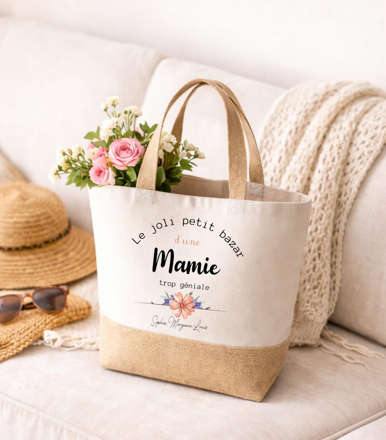 May include: A canvas tote bag with a natural jute bottom and handles. The bag features the text "Le joli petit bazar d'une Mamie trop g&eacute;niale" with a floral design. The bag is filled with pink roses and white flowers.