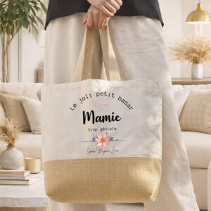 May include: A beige tote bag with a natural jute bottom and handles. The bag features the text "Le joli petit bazar d'une Mamie trop g&eacute;niale" in black script, with a floral design. The bag is held by a person.