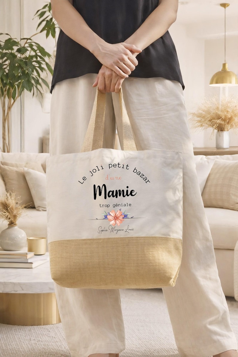 May include: A beige tote bag with a natural jute bottom and handles. The bag features the text "Le joli petit bazar d'une Mamie trop g&eacute;niale" in black script, with a floral design. The bag is held by a person.