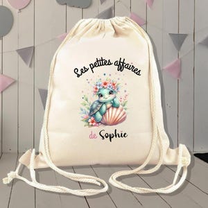 Könnte beinhalten: Eine weiße Kordeltasche mit einer niedlichen Comic-Schildkröte, die eine Blumenkrone trägt und eine Muschel hält. Die Tasche ist mit dem Text "Les petites affaires de Sophie" bedruckt.