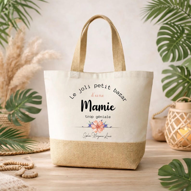 May include: A beige tote bag with a natural jute bottom and handles. The bag features the text "Le joli petit bazar d'une Mamie trop g&eacute;niale" in black script, with a floral design and a name below.