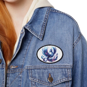 May include: A denim jacket with a patch featuring a colorful phoenix design. The patch is oval-shaped with a black border, showcasing a blue, purple, and white phoenix. The jacket has a classic design with buttoned pockets and a collar.