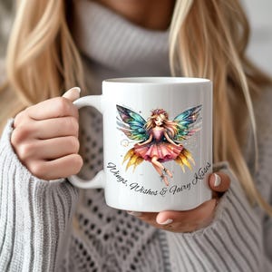 May include: White ceramic mug featuring a watercolor illustration of a fairy with colorful wings and a pink dress. The mug has the text "Wings, Wishes & Fairy Kisses" in a script font. The mug is held by a person.
