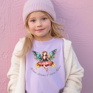 May include: A young girl wearing a light purple t-shirt with a fairy graphic and the text "Wings, Wishes & Fairy Kisses." She is also wearing a light purple beanie and a white cardigan. The word "Orchid" is printed at the bottom left.