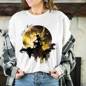 May include: White t-shirt with a graphic of a fairy sitting on a crescent moon with the text "Fairy Magic" above the moon.