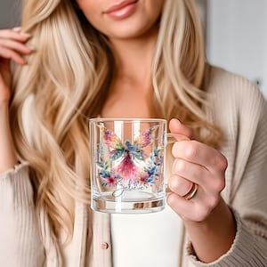 Puede incluir: Una taza de vidrio transparente con un diseño de hada colorida y la palabra "Believe". El hada tiene alas iridiscentes y está rodeada de detalles florales. La taza tiene un asa. El diseño es en tonos azules, rosas y morados.