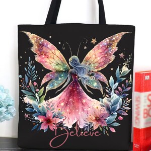 May include: Black tote bag featuring a watercolor illustration of a fairy with butterfly wings, surrounded by flowers and foliage. The word "Believe" is written in cursive. The bag has black handles.