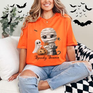 May include: Orange t-shirt with a cartoon mummy, ghost, and black cat holding a pumpkin. The text "Spooky Season" is printed on the shirt.
