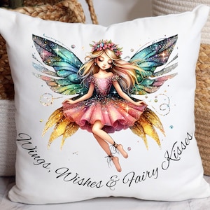 Puede incluir: Cojín cuadrado blanco con una ilustración de hada. El hada tiene alas coloridas y un vestido rosa, adornado con una corona de flores. El texto "Wings, Wishes & Fairy Kisses" está escrito en cursiva en el cojín.