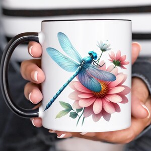 May include: White ceramic mug with a black handle and rim, featuring a detailed illustration of a blue dragonfly with pink and white flowers. The dragonfly has intricate wing patterns and is set against a white background.