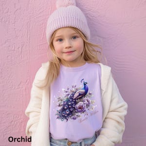 May include: A young girl wearing a light purple t-shirt with a peacock design. The peacock is depicted with blue, purple, and white feathers, surrounded by flowers. The text "Beauty Within" is below the peacock. She is also wearing a light purple beanie and a white cardigan.