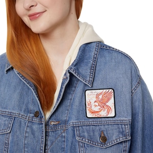 Könnte beinhalten: Hellblaue Jeansjacke mit einem quadratischen Aufnäher, der einen Phönix in Rot- und Orangetönen zeigt. Der Aufnäher trägt den Text "Fly above Earth below". Die Jacke hat einen Knopfverschluss.