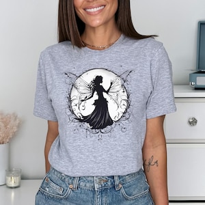 Puede incluir: Una camiseta gris con una silueta negra de un hada con alas en un círculo de enredaderas y estrellas.