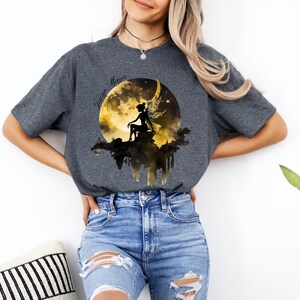 May include: A dark gray t-shirt with a graphic of a fairy sitting on a crescent moon with the text "Fairy Magic" above the moon.