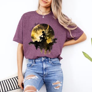 May include: A purple t-shirt with a graphic of a fairy sitting on a crescent moon with the words "Fairy Magic" in a black script font.