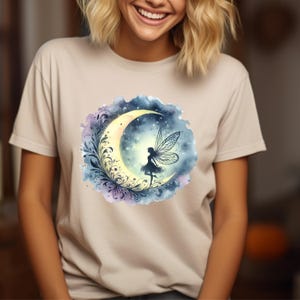May include: A light beige t-shirt featuring a watercolour design of a fairy standing on a crescent moon. The moon is surrounded by a floral design and stars. The background is a blue and purple watercolour wash.