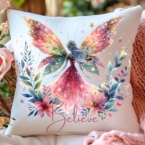 Puede incluir: Un cojín cuadrado decorativo con un diseño de hada, que muestra un hada con alas vibrantes y un vestido en tonos rosas y rojos. Elementos florales en rosa, azul y morado rodean el hada. La palabra "Believe" está escrita en cursiva.
