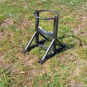 May include: A black metal stand with a circular top and a V-shaped support. The stand has a square base and is designed to hold a cylindrical object. The stand is made of welded metal and is sitting on grass.