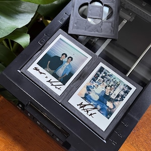May include: A black scanner with two Polaroid photos and a film holder. The photos feature images of people with signatures. The film holder is square with a series of openings. The scanner is on a wooden surface.