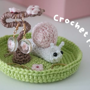 May include: A crocheted snail with a pink shell and black eyes sits on a green crocheted dish. The snail is holding a small branch with pink flowers and two pairs of earrings. The dish has two rings and a pink flower on it. The text "Crochet Pattern" is written in white on a pink background.