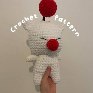 May include: White crocheted amigurumi toy with a red pom-pom on top and a red nose. The toy has a stitched-on smile and is holding its arms out. The text "Crochet Pattern" is written in white on a white background.
