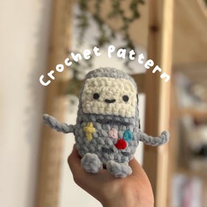 Puede incluir: Juguete de peluche gris tejido a crochet con forma de consola de videojuegos con botones de colores y una estrella amarilla. El juguete tiene una cara blanca con una boca sonriente y dos ojos negros. El texto "Crochet pattern" está escrito en blanco sobre un fondo borroso.