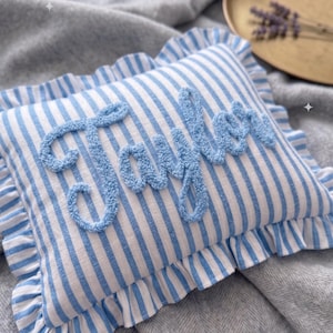 Può includere: Un cuscino rettangolare a righe blu e bianche con bordo a balze. La parola "Taylor" è ricamata in azzurro sul davanti. Il cuscino è appoggiato su una coperta grigia.