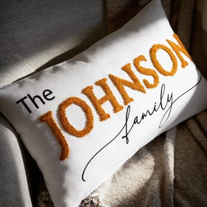 May include: White rectangular pillow with the text "The JOHNSON Family". The word "JOHNSON" is in orange, textured, raised letters. The word "The" is in black, and "Family" is in a black script font.