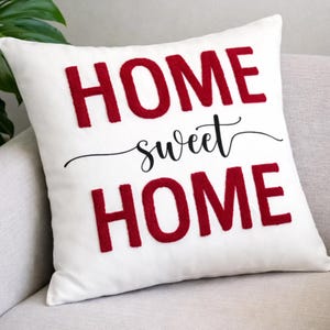 May include: A white square pillow with the words "HOME sweet HOME" in red and black. The letters are raised, giving a textured appearance. The pillow is on a light gray surface.
