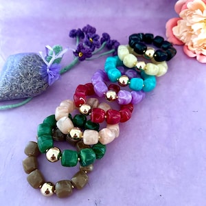 Op de afbeelding: Een verzameling kleurrijke armbanden met kralen in verschillende tinten, waaronder groen, rood, paars en zwart. Elke armband heeft goudkleurige accentkralen. De armbanden liggen op een lavendelkleurig oppervlak, met een lavendelzakje en bloemen op de achtergrond.